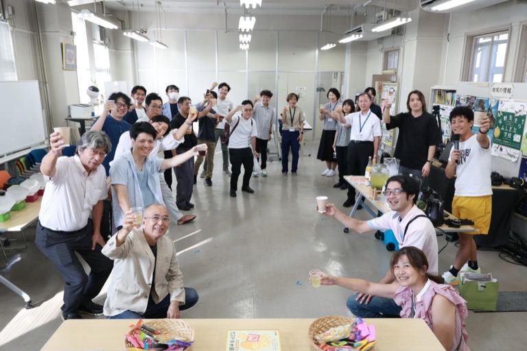 懇親会で25名ほどの参加者が飲み物を持って笑顔であがりポーズを決めている