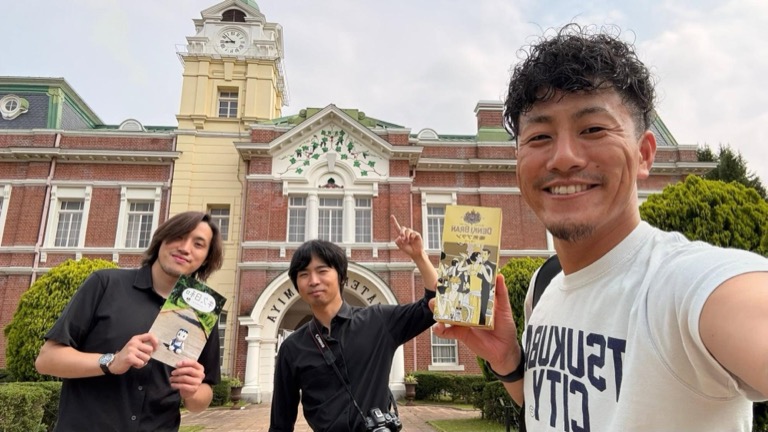 時計台付きの歴史的建造物前で、堀下さん・川原さん・友也さんが商品パンフレットを持って記念撮影している